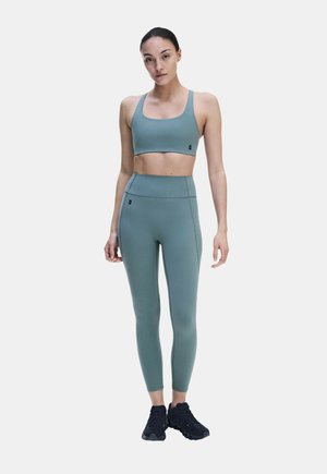 Frau trägt hellblaue Leggings mit hoher Taille und passenden Sport-BH sowie schwarze Sportschuhe, steht vor einfachem Hintergrund.