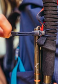 Bâton de trekking noir et orange avec une poignée en caoutchouc, fixé par une attache et un cordon élastique, sur fond de sac à dos bleu.