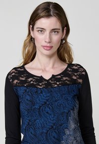 Haut noir à manches longues avec un empiècement en dentelle, présentant des motifs paisley bleus complexes sur le corps et des détails de décolleté boutonnés.