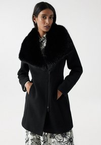 Manteau en laine noir avec un grand col en fausse fourrure noire douce, fermeture éclair et poches latérales. Le modèle porte une robe à motifs floraux en dessous.