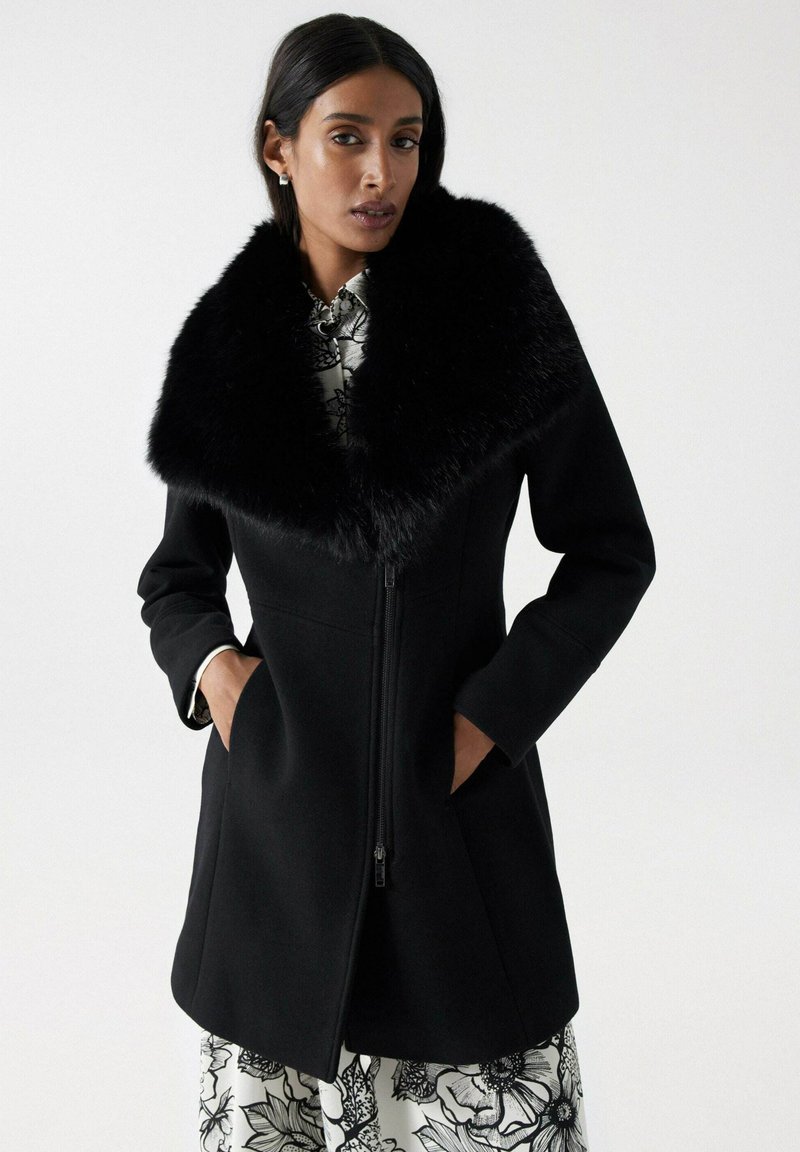 Manteau en laine noir avec un grand col en fausse fourrure noire douce, fermeture éclair et poches latérales. Le modèle porte une robe à motifs floraux en dessous.