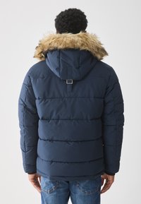 Chaqueta de plumas azul marino con capucha forrada de piel, que presenta secciones acolchadas horizontales y un detalle de correa trasera para ajuste.