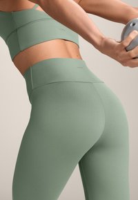 Svijetlozelena aktivna odjeća koja se sastoji od prilagođenog sportskog grudnjaka jednostavnog dizajna i visokih tajica glatke teksture.