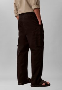 Homme portant un pantalon cargo noir avec ceinture élastique et poches latérales, associé à une chemise beige et des sandales noires, debout de profil.