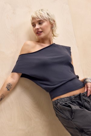 Blondínka sa opiera o svetlú drevenú stenu, nosí tmavý top s odhalenými ramenami, voľné tmavé nohavice, náramky a tetovania psov na ľavej ruke.