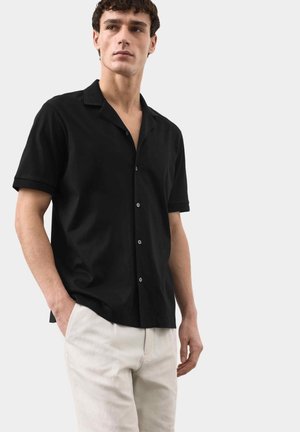 Ung mand iført en sort kortærmet skjorte med knapper og lysebeige bukser, stående med den ene hånd i lommen mod en ensfarvet baggrund.