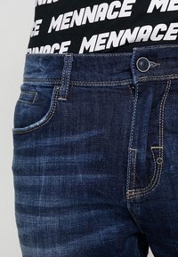 Dark blue denim jeans with a slightly faded finish. Features include a five-pocket design, gold stitching, and a metallic button closure.