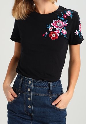 Femme portant un haut noir à manches courtes avec une broderie florale colorée sur l'épaule et la manche, associée à une jupe en denim bleu boutonnée sur le devant.