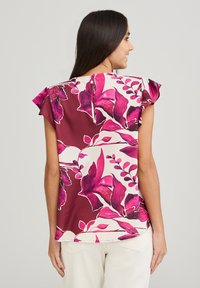 Blusa floreale con maniche corte a volant, caratterizzata da una combinazione di colori marrone e crema, con vivaci motivi floreali rosa. Chiusura con bottoni sulla schiena.