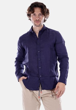 Uomo con capelli ricci castani, indossa una camicia blu navy con bottoni e pantaloni beige, in piedi con una mano in tasca.