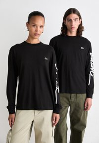 Schwarze Langarmshirts mit einem kleinen grünen Emblem, weißem Text auf den Ärmeln, kombiniert mit beigen Cargo-Hosen und olivgrünen Hosen.