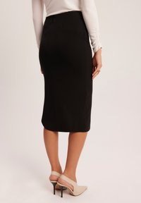 Czarna spódnica ołówkowa o gładkiej fakturze, średniej długości, z delikatnymi detalami szwów, zestawiona z beżowymi sandałami na otwartym palcu na neutralnym tle.