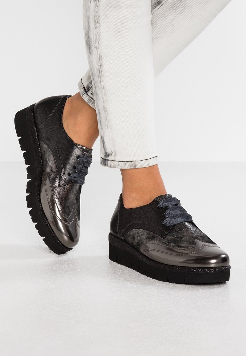 Chaussures à lacets noires et métalliques avec un dessus texturé, une semelle épaisse et des lacets bleus, sur un fond en denim clair.