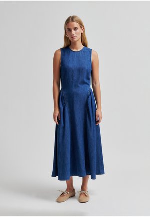 Ung kvinne står iført ermeløs blå denim midi-kjole med rynkede sidelommer og beige snøresko foran ensfarget bakgrunn.