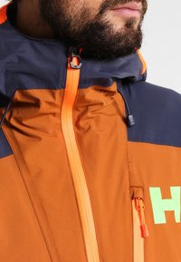 Veste à capuche avec panneaux orange et bleu marine, dotée d'une fermeture éclair frontale, d'accents réfléchissants et d'un logo proéminent en vert vif.