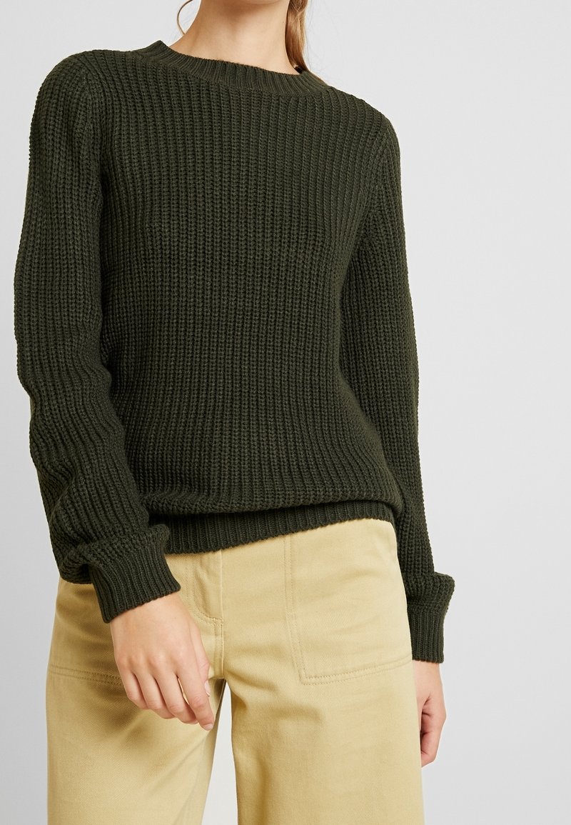 Personne portant un pull en grosse maille vert foncé et un pantalon beige, debout devant un fond clair uni.