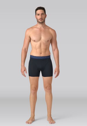 Homme torse nu, debout pieds nus, portant un boxer noir avec une ceinture bleu marine portant l'inscription "Muchachomalo" sur un fond gris.