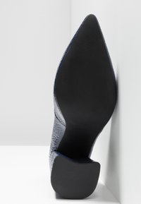 Chaussure à talon aiguille marine avec un bout pointu, une tige texturée et une semelle noire lisse. Le talon est fin et angulaire, ce qui renforce le design épuré.