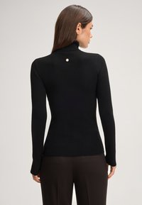 Zwarte geribbelde coltrui met lange mouwen, voorzien van een kleine metalen accenten op de rug. Glad oppervlak en aangesloten ontwerp.