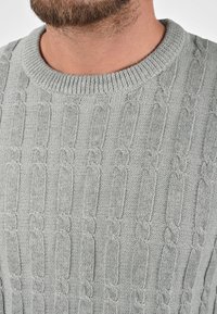 Pull en tricot gris clair avec un motif texturé comprenant des câbles en relief et un col rond côtelé, mettant en valeur un tissu doux et confortable.