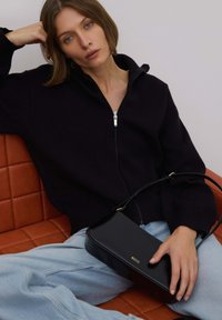 Black zip-up sweater with ribbed cuffs, paired with light blue jeans. A sleek black handbag with a gold logo rests on the lap, on an orange couch.