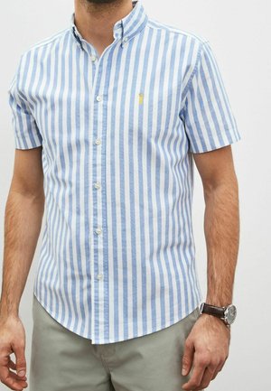 Homme portant une chemise à manches courtes à rayures verticales bleues et blanches avec un petit logo brodé jaune sur la poitrine et un pantalon gris.