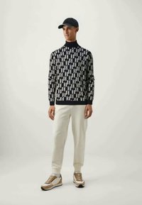Mannen navy turtleneck trui met wit geometrisch patroon, crèmekleurige broeken en lichte sneakers met gouden accenten; baseballpet op het hoofd.