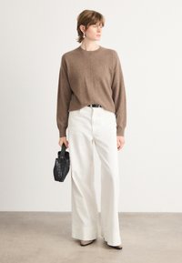 Καφέ πλεκτό πουλόβερ με μακριά μανίκια, συνδυασμένο με λευκά παντελόνια με φαρδιά γραμμή. Το μοντέλο κρατά μια μικρή μαύρη υφασμάτινη τσάντα. Απλός, μοντέρνος σχεδιασμός.