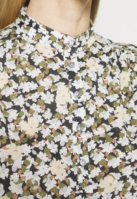 Bloemenblouse van lichtgewicht stof met een zwarte achtergrond en witte, groene en perzikkleurige bloemen, een hoge kraag en doorzichtige knopen.