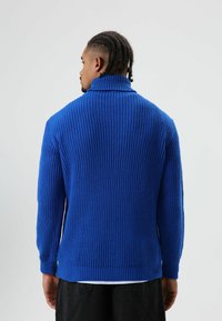 Dikgebreide blauwe trui met een hoge kraag en een geribbelde structuur. Lange mouwen en een iets verlengde rugzoom. Gecombineerd met een donkere broek.