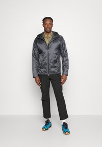 Graue Puffjacke mit Kapuze, glänzende Textur, Reißverschluss vorne, kombiniert mit olivgrünem Hemd, schwarzen Hosen und blauen Sportschuhen.