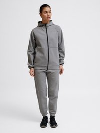 Frau steht und trägt einen grauen Kapuzenpullover mit Reißverschluss und passende Jogginghosen mit schwarzen Sneakers vor einem schlichten Hintergrund.