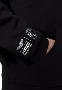 Czarna bluza z białymi logo: Aston Martin, Aramco i emblemat języka Rolling Stones na rękawie. Miękka tkanina z dodatkową kieszonką.