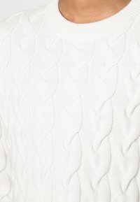 Vit stickad tröja med kabelmönster som har rund halsringning och strukturerat mönster på tyget. Mjuk, kraftig stickning med upphöjda, vågiga mönster.