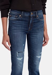 Jeans in denim blu scuro con design aderente, caratterizzati da dettagli logori tra cui strappi alle ginocchia e leggere scoloriture in tutto il tessuto.
