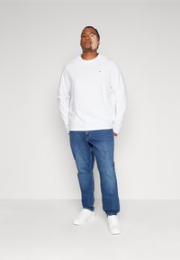 Hvit langermet skjorte, denimjeans og hvite joggesko. Har en liten logo på brystet. Rett passform og avslappet design.