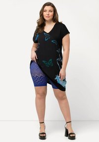 Zwarte tuniek met korte mouwen, versierd met blauwe vlinderprint, gecombineerd met blauwe zigzagpatroon shorts. De outfit wordt compleet gemaakt met zwarte sleehakken.