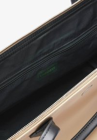 Wnętrze torby Lacoste z czarną tkaniną wykończeniową. Posiada jedną kieszeń na zamek błyskawiczny, srebrny suwak, beżowy zewnętrzny kolor i czarne skórzane uchwyty.