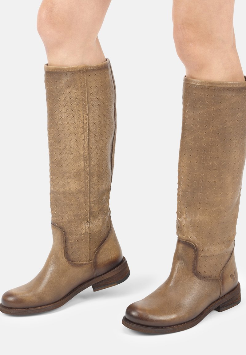 Bottes en cuir marron montant jusqu'aux genoux avec un motif croisé subtil, bouts arrondis, talons bas et semelles lisses, portées sur des jambes nues sur un fond blanc.