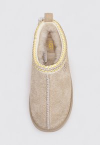 Chausson UGG beige à enfiler avec des coutures décoratives jaunes, doublure en polaire douce et languette à l'arrière.