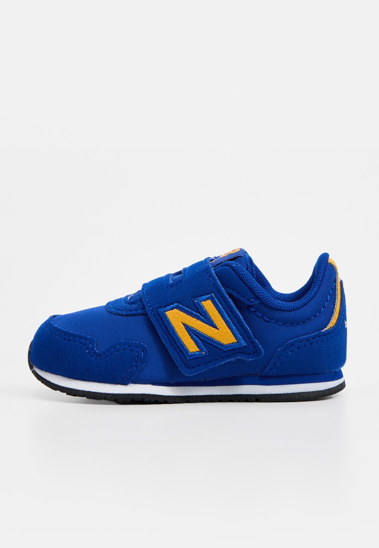 Chaussure de sport bleue avec une sangle Velcro, arborant un logo "N" jaune. Matériau lisse, bout arrondi, semelle noire avec bord blanc et accents de couture.