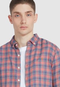 Młody mężczyzna z krótkimi włosami, ubrany w koszulę w kratę w czerwono-niebieskie wzory na białym t-shircie, stojący przodem na jednolitym tle.