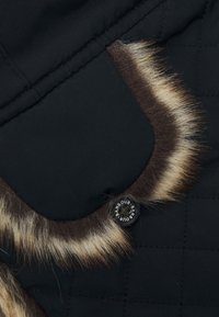 Czarna pikowana tkanina z futrzanym wykończeniem; posiada okrągły czarny guzik z logo "Barbour". Miękka tekstura, kontrastujące kolory w akcentach.