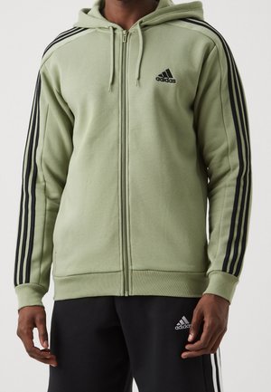 Mężczyzna w jasnym zielonym bluzie z kapturem Adidas na zamek, z czarnymi trzema paskami na rękawach i czarnych spodniach dresowych Adidas z logo na udzie.