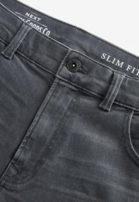 Jeans slim gris foncé au niveau de la taille avec fermeture par bouton, passants de ceinture et texte de marque discret sur la ceinture intérieure.