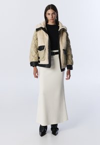 Femme portant une veste beige matelassée et doublée de fourrure, sur un haut noir, une longue jupe blanche avec une ceinture noire, et des bottes noires à bout pointu, se tenant devant un fond blanc.