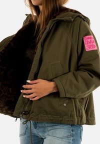 Donna che indossa una giacca verde oliva con un patch rosa che dice "Born a Bad Seed", con foderatura in pelliccia marrone e jeans blu chiaro.
