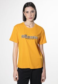 Gul bomulds T-shirt med korte ærmer, rund halsudskæring og #Bossin grafik i sort og sølv skrift på fronten.