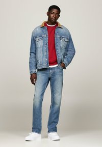 Chaqueta de mezclilla azul claro con cuello marrón, sudadera roja y jeans azules claros. Zapatillas blancas completan el atuendo casual.
