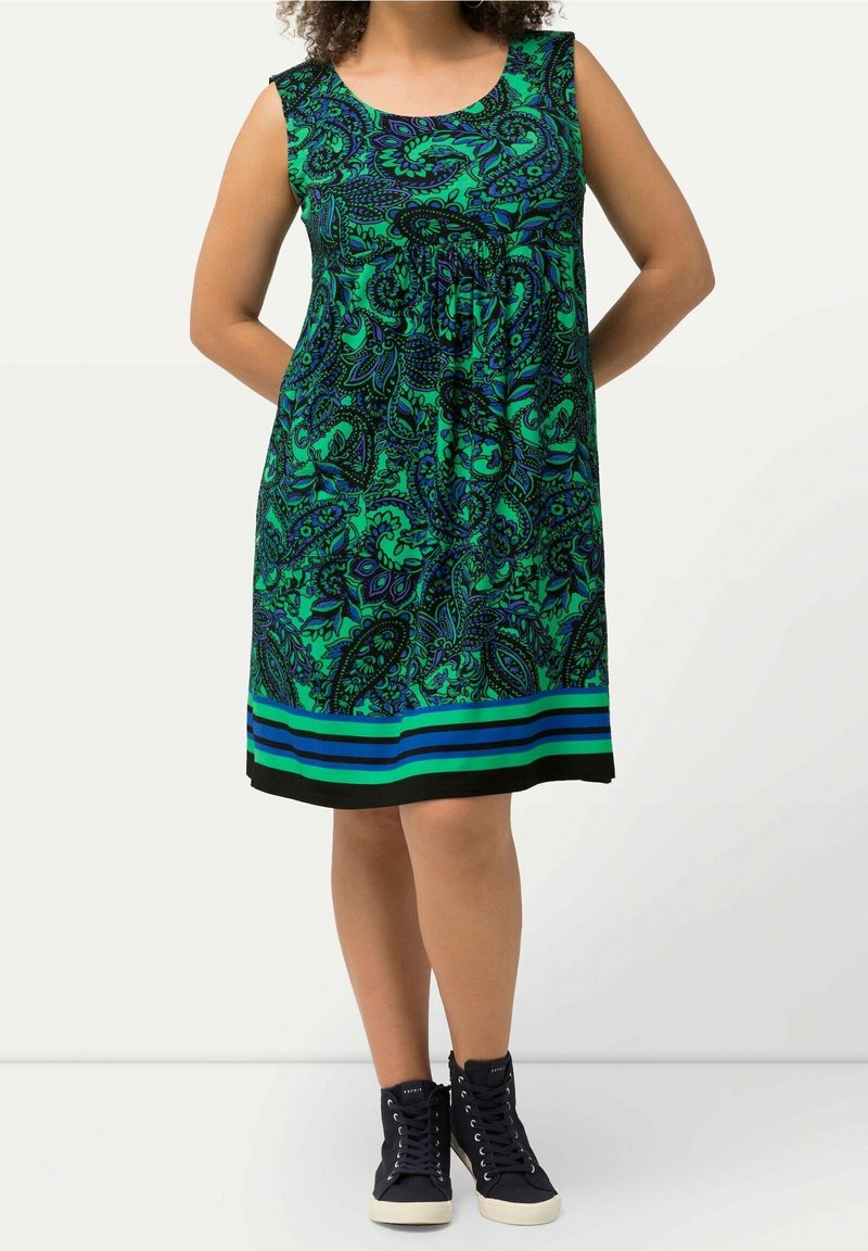 Femme portant une robe verte sans manches avec un imprimé cachemire bleu et noir et des baskets noires, debout, les mains derrière le dos.